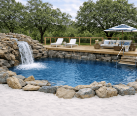 Natural swimming hole with a small cascading waterfall flowing into a clear pool, bordered by a wooden deck with chairs on one side and a sandy edge on the other, surrounded by lush greenery